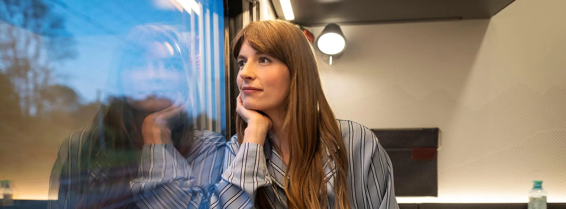 Eine junge Dame mit langen Haaren im Schlafanzug, die in einem Abteil des Nachtzuges Nightjet sitzt und aus dem Fenster schaut. 