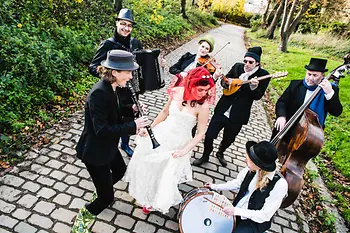 Jiddische Musikband spielt im Grünen auf einem gepflasterten Weg, dazu tanzt eine Braut.