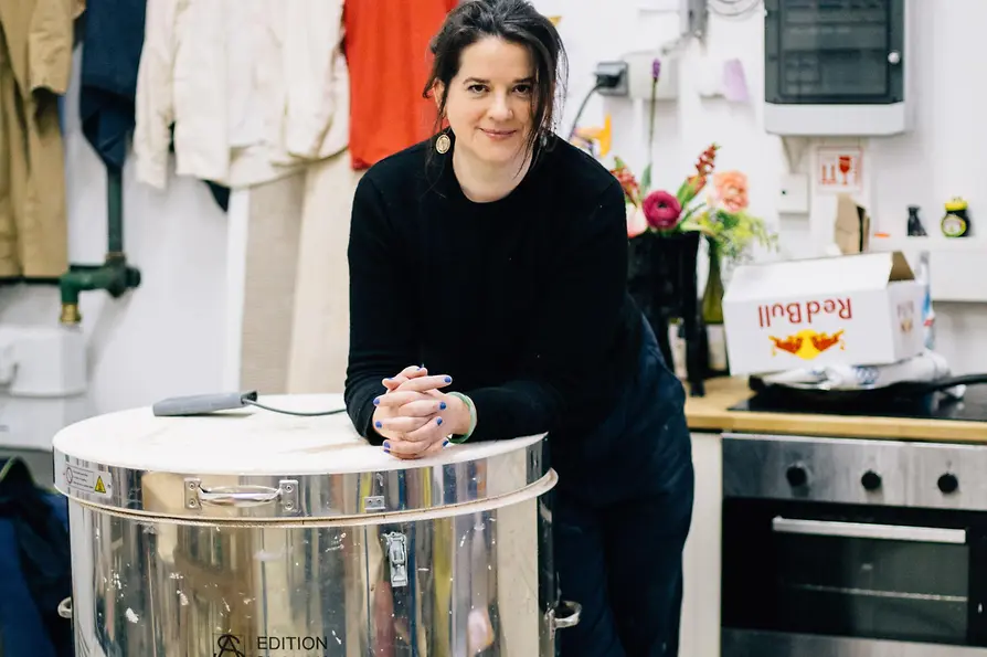 Artist in her studio leaning against a ceramic kiln, surrounded by tools and clothing.