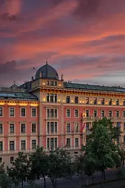 The historic façade of the Anantara Palais Hansen Vienna glows warmly at sunset, with a dramatic sky of red and purple clouds above the grand building lined with trees.