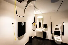 Innenraum einer barrierefreien Toilette mit deckenmontiertem Hebesystem und hängender Fernbedienung, einem Waschbecken mit Spiegel sowie Stützgriffen neben dem WC.