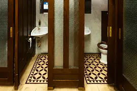 Innenansicht einer historischen öffentlichen Toilette mit dunklen Holztüren, Milchglasfenstern, gemustertem Fliesenboden und Vintage-Waschbecken vor Marmortafeln.