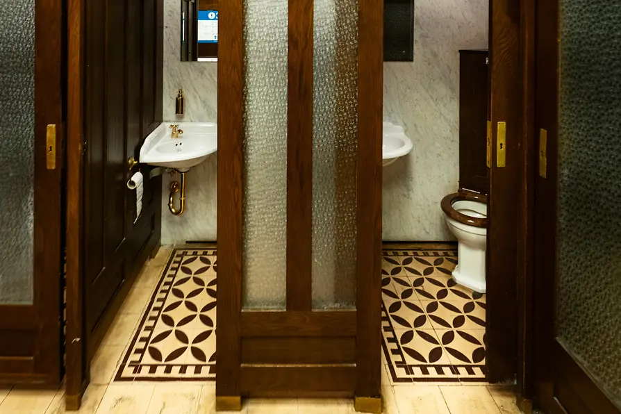 An interior view of a historic public restroom with dark wooden doors, frosted glass panels, patterned tile flooring, and vintage sinks with marble walls.