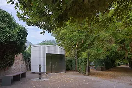 Ein modernes öffentliches WC-Gebäude mit metallischer Fassade entlang eines baumbestandenen Weges im Augarten. Laub liegt teilweise am Boden, Bänke und Hecken rahmen die ruhige Parksituation ein.