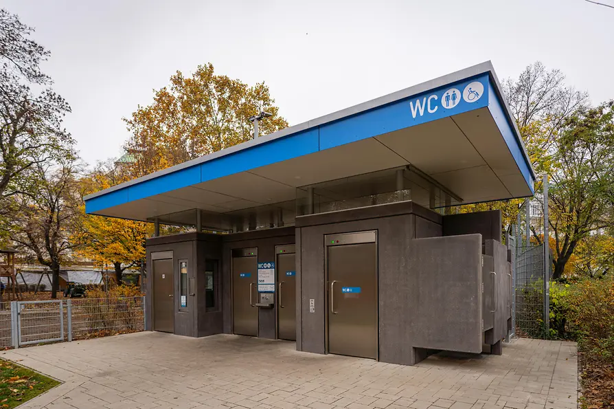 Eine moderne öffentliche WC-Anlage mit blauem Dachabschluss, dunkelgrauen Wänden, Edelstahltüren und barrierefreien Hinweisschildern, umgeben von Herbstlaub.
