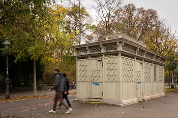 Ein historischer, hellgrauer WC-Pavillon mit dekorativen Gitterelementen und bunten Glasdetails, umgeben von Herbstbäumen, während Fußgänger vorbeigehen.