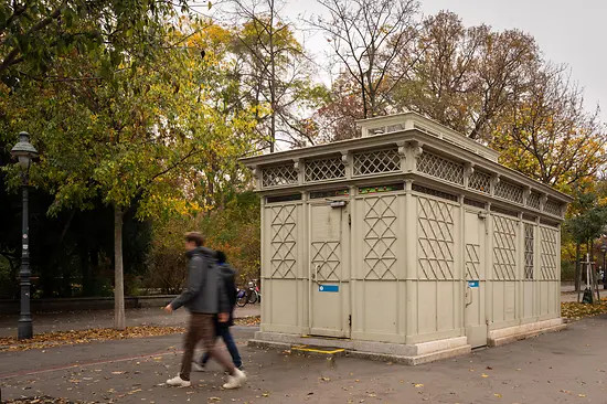 Ein historischer, hellgrauer WC-Pavillon mit dekorativen Gitterelementen und bunten Glasdetails, umgeben von Herbstbäumen, während Fußgänger vorbeigehen.