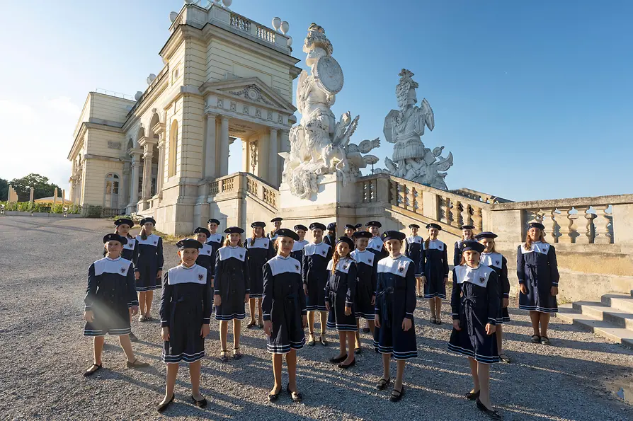 Fetele din corul vienez, în uniforme, stau în fața Gloriettei din parcul Palatului Schönbrunn, cu arhitectură istorică în fundal.