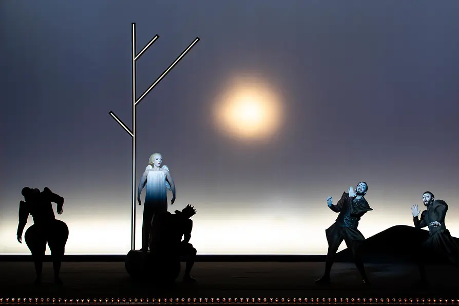 Escena de ópera con varios intérpretes ante una escenografía minimalista con una fuente de luz intensa.