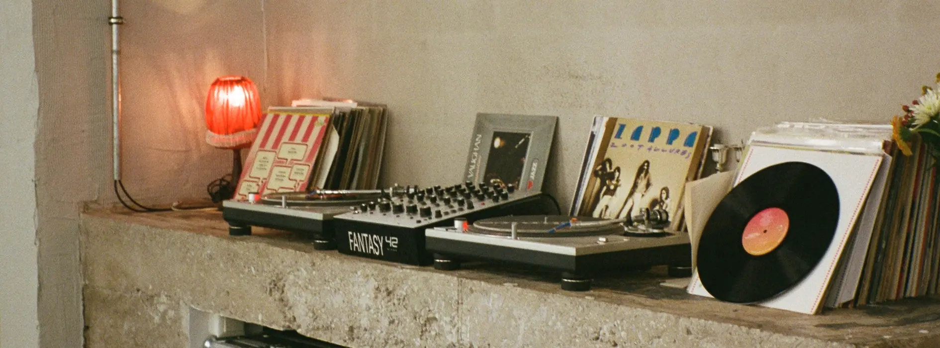 Turntables and a mixer sit on a concrete shelf next to stacked vinyl records; a small red lamp glows by the window.
