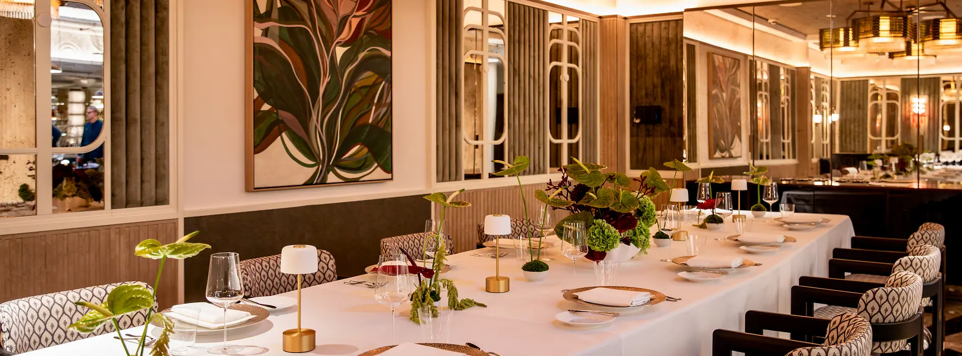 A long elegantly set table with floral arrangements and small lamps in a refined dining room with mirrors and chandeliers.