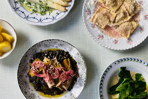 Several plates of seasonal dishes are arranged on a light tablecloth, including white asparagus, filled pasta, and leafy greens.