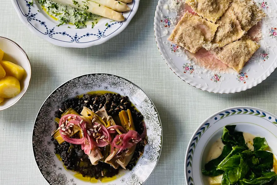 Several plates of seasonal dishes are arranged on a light tablecloth, including white asparagus, filled pasta, and leafy greens.