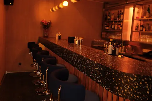 A long terrazzo bar counter with blue bar stools and warm Edison bulbs hanging from the ceiling.