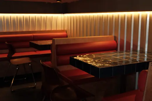 A dimly lit bar area with red upholstered booths, mosaic tile tables, and a warm light strip along the wall.