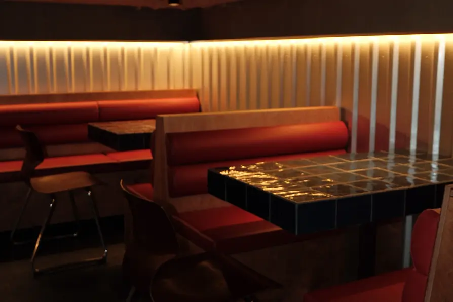A dimly lit bar area with red upholstered booths, mosaic tile tables, and a warm light strip along the wall.