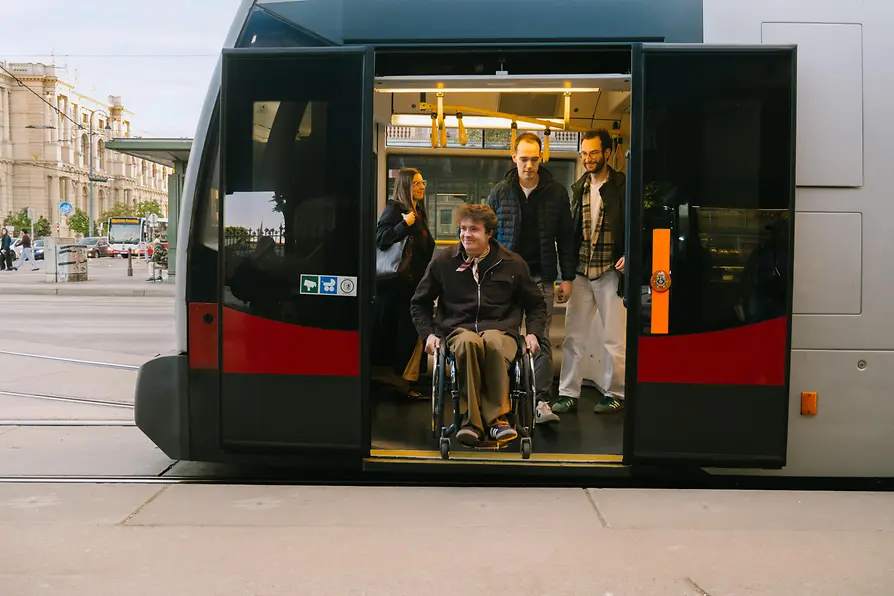 A man using a wheelchair is exiting a Wiener Linien tram while other passengers remain inside the vehicle. The image shows accessible mobility on public transport.