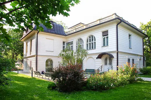 Klimt Villa inmitten eines weitläufigen Gartens mit Blumenbeeten und Bäumen.