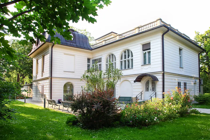 Klimt Villa inmitten eines weitläufigen Gartens mit Blumenbeeten und Bäumen.