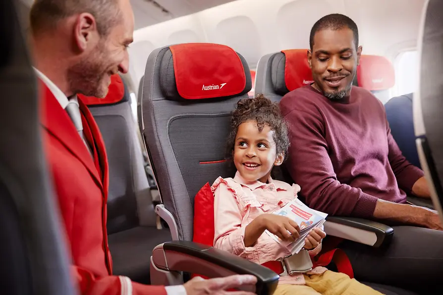 Ein Mann und ein Kind sitzen in der Economy Class eines Austrian-Airlines-Flugs, während ein Flugbegleiter in roter Uniform mit ihnen spricht.