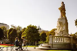 Két ember ül a Burggartenban a napon Mozart szobra előtt