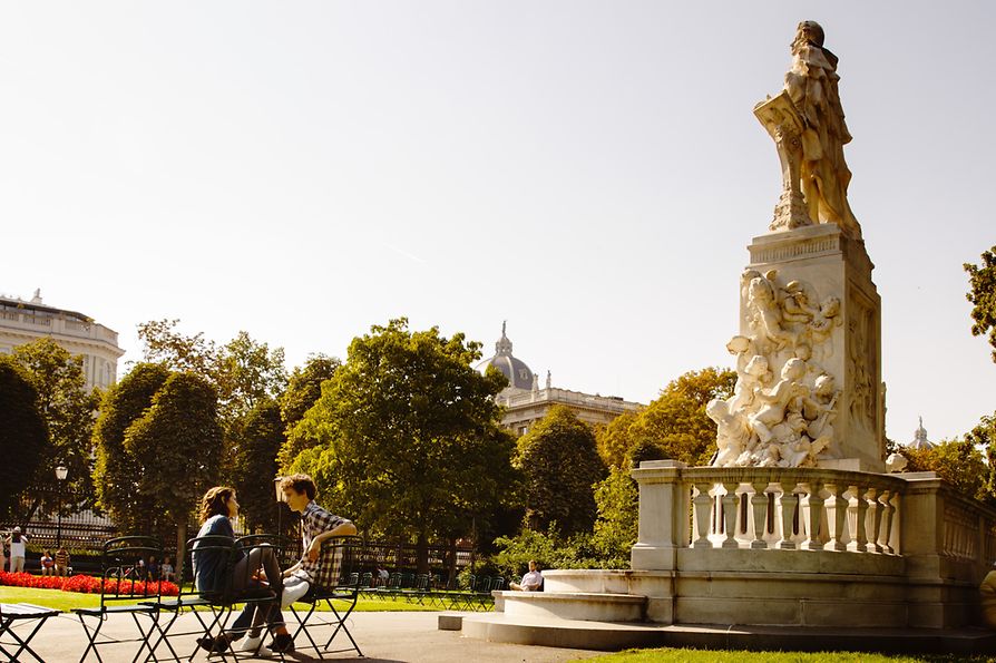 Doi oameni stau la soare în Parcul Burggarten, în faţa statuii lui Mozart