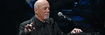 Billy Joel apoyado en el piano, con el micrófono delante.