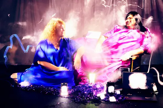 Queere Performance AFTERLIVES der Kollektive Henrike Iglesias & Miss Theatre. Zwei Akteur:innen in wallenden Gewändern auf der Bühne sitzend.