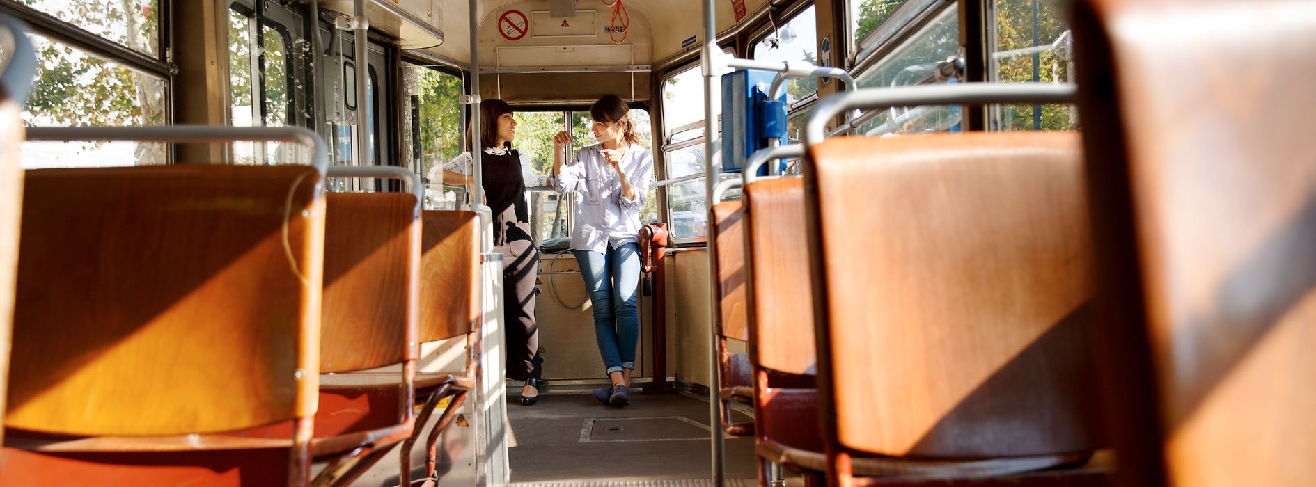 Zwei Frauen stehen in einer Straßenbahn und unterhalten sich, im Vordergrund leere Sitze