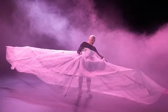 A female dancer in an oversized, sheer hoop skirt bathed in pink light.