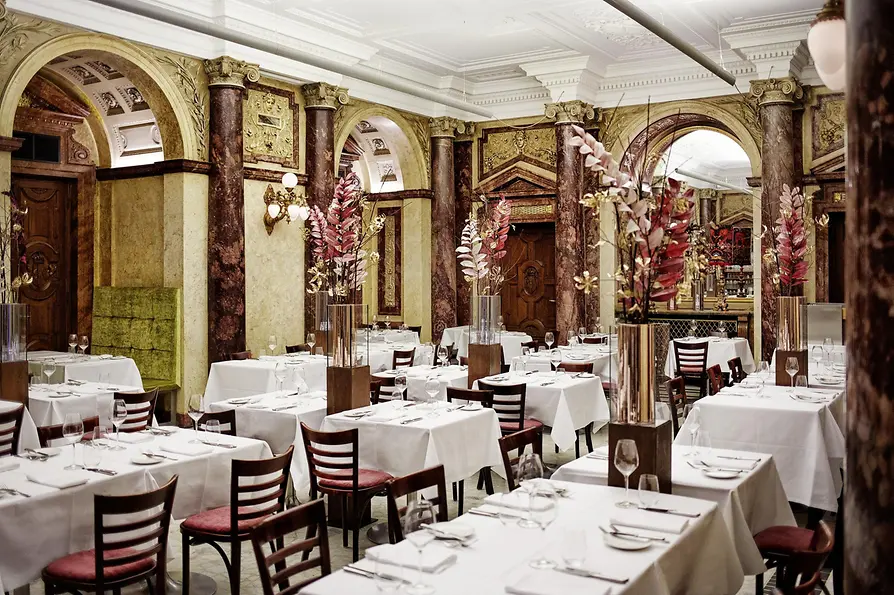 Prunkvoller Speisesaal mit Marmorsäulen, Rundbögen, hohen Blumenarrangements und weiß gedeckten Tischen in klassischem Ambiente.