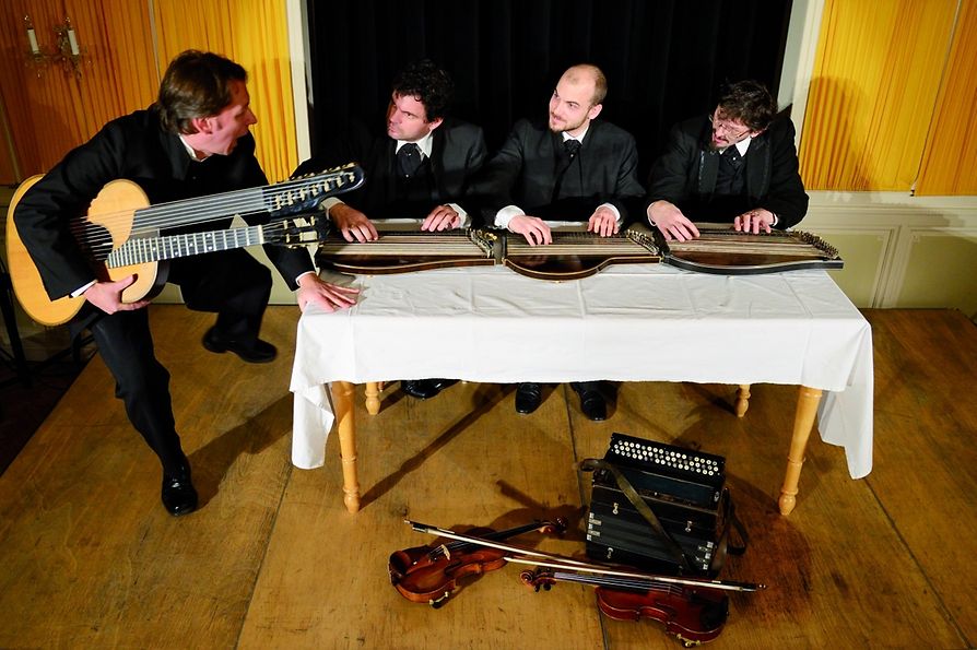 Sujet Schrammelmontag: Musiker an einem Tisch mit Instrumenten, in einer holzvertäfelten Gaststube