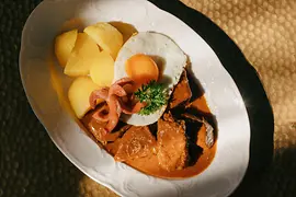 Un plato con un gulasch con patatas cocidas, huevo frito y una salchicha