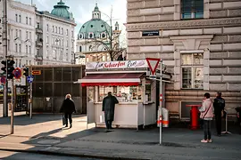 A sausage stand, people in front of it, in the background the dome of the Karlskirche (Church of St. Charles)