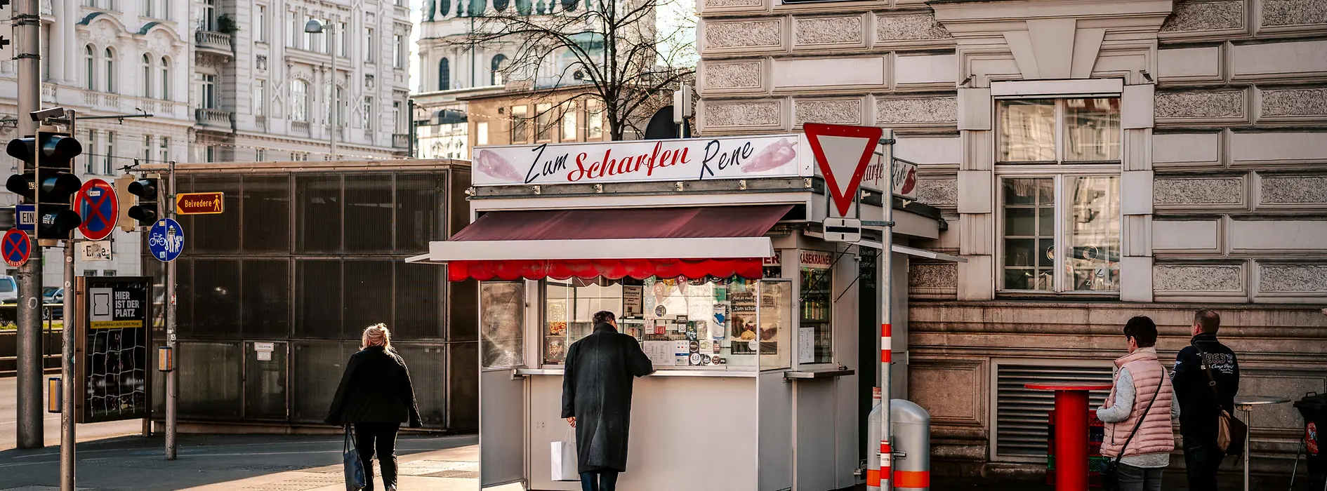 A sausage stand, people in front of it, in the background the dome of the Karlskirche (Church of St. Charles)