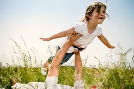Little girl who is lifted up by a man lying on grass while she pretends to fly.