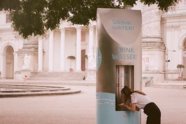 Frau trinkt an einem Trinkbrunnen, im Hintergrund die Karlskirche