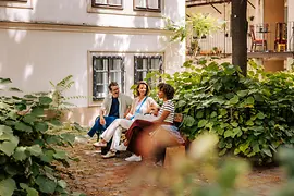 Two women and a man on a bench in a leafy courtyard