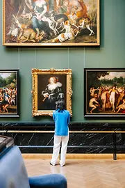 Woman with dark hair a blue shirt and white pants looks at a historic artwork in a golden frame at the Art History Museum in Vienna