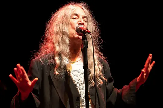 Patti Smith canta de pie en un escenario oscuro al micrófono.