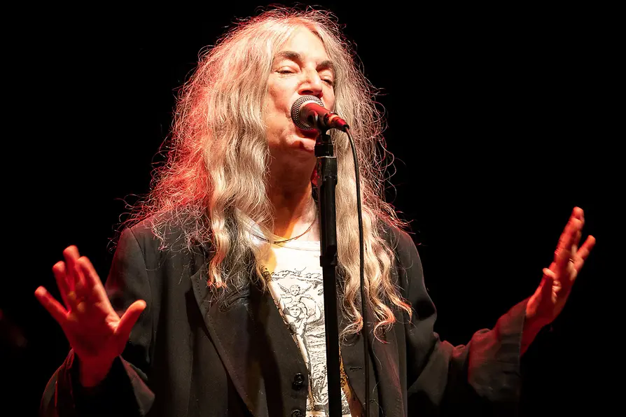 Patti Smith canta de pie en un escenario oscuro al micrófono.