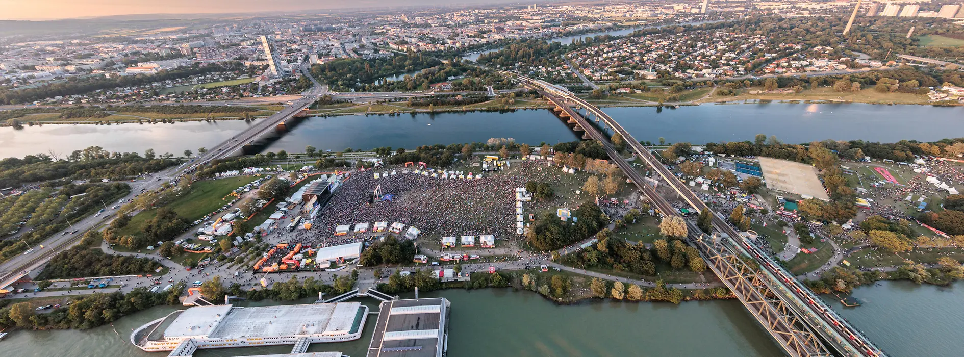Luftaufnahme mit Blick auf die Donauinsel, auf der das Open Air Donauinselfest gerade stattfindet, alles ist voller Menschen, Bühnen und Zelte.
