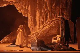 Theatre scene in orange lighting against a rocky backdrop, several performers in different costumes on stage.