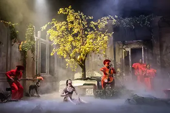 Scène théâtrale avec des musiciens en vêtements rouges, une danseuse assise au sol et un arbre à feuilles caduques sur un lit double. 