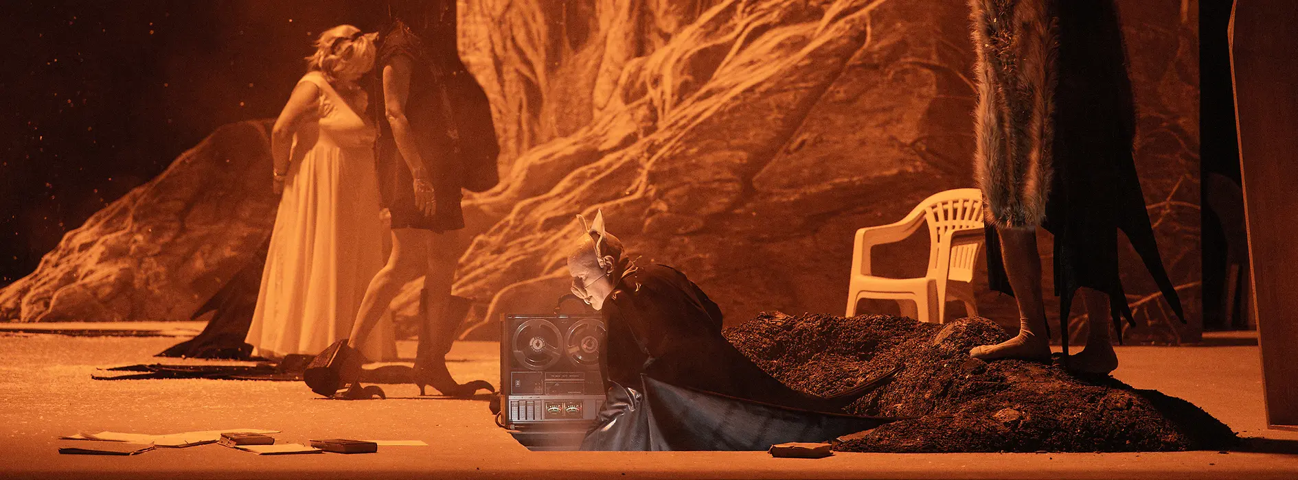 Theatre scene in orange lighting against a rocky backdrop, several performers in different costumes on stage.