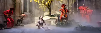 Scène théâtrale avec des musiciens en vêtements rouges, une danseuse assise au sol et un arbre à feuilles caduques sur un lit double. 
