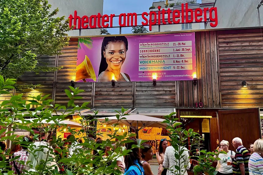 Außenansicht des \"Theater am Spittelberg\" mit rotem Schriftzug. Ein großes Plakat mit dem Foto einer lächelnden Frau und Informationen zu Veranstaltungen hängt an der Holzfassade. Vor dem Eingang stehen Menschen unter Sonnenschirmen und unterhalten sich.