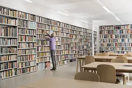 Bibliothek des queeren Zentrums Qwien mit vollen Bücherregalen, Tischen, Stühlen und einem Bibliothekar, der gerade ein Buch zurück in das Regal stellt