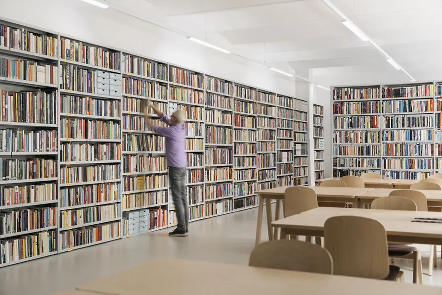 Bibliothèque du centre queer Qwien avec des étagères remplies de livres, des tables, des chaises et un bibliothécaire remettant un livre sur une étagère