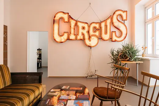 Espace d’accueil du centre queer Qwien avec un canapé, une table et des chaises, et une grande enseigne lumineuse de la série de soirées Circus sur le mur